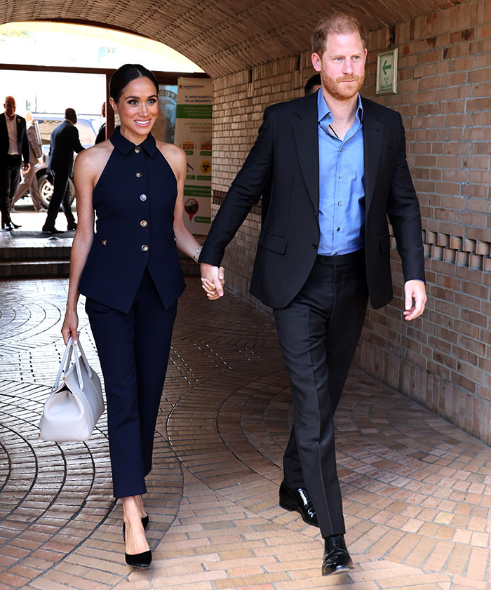 Royal couple holding hands in a formal setting, both in navy and black outfits, related to Hollywood news.