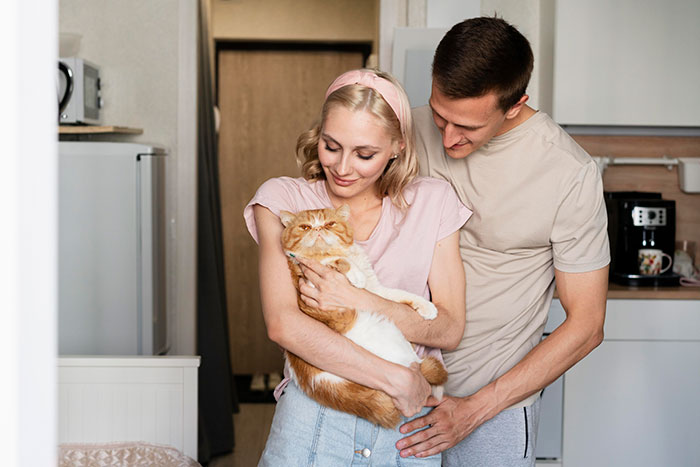 A smiling couple in a cozy kitchen, with the woman holding an orange cat, illustrating a lighthearted moment in relationships.
