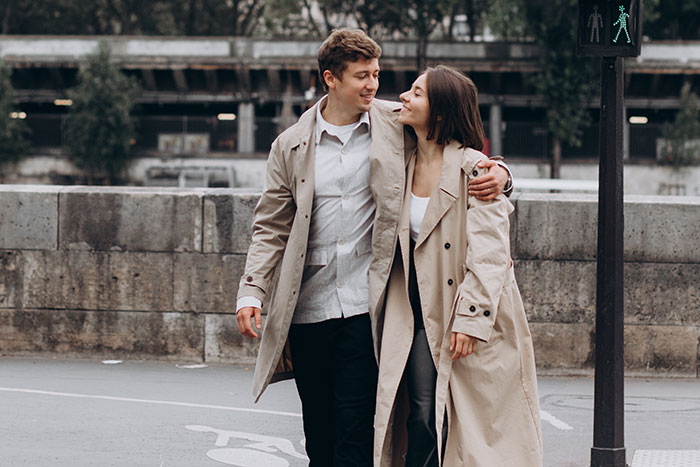 A couple walking together in trench coats, smiling at each other, outdoors.