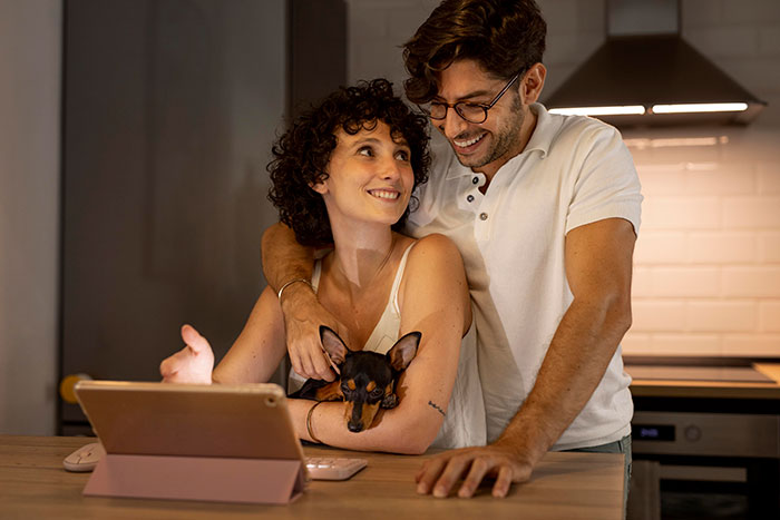 Couple smiling in kitchen, man embracing woman, with a brown dog and a tablet on the table.