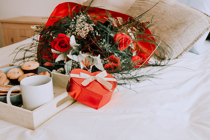 Bouquet of red roses and a heart-shaped gift box on a bed, alongside a tray with a mug and pastries, theme of lies men tell.