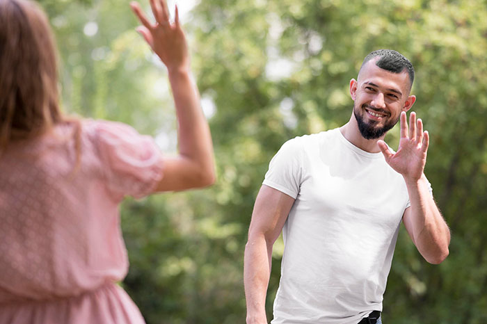 Man in white shirt smiling and waving in park, engaging in conversation with partner.