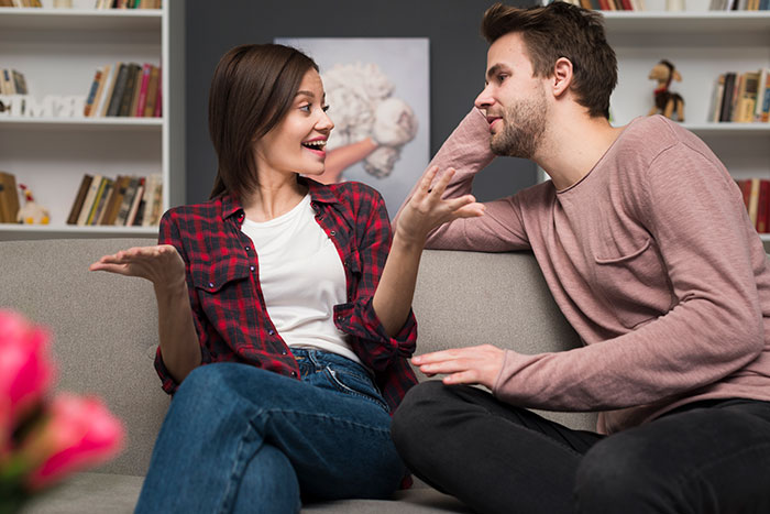 A couple sitting on a couch, the man leaning towards the woman, both smiling, in a cozy living room setting.
