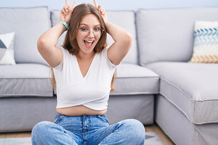 Woman sitting on the floor, playfully making bunny ears gesture, wearing glasses and a white shirt, smiling in a casual setting.