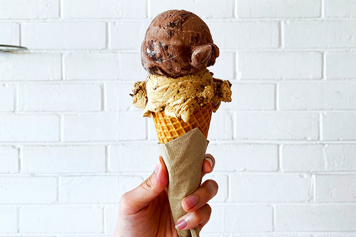 Hand holding a chocolate and vanilla ice cream cone against a white brick wall.