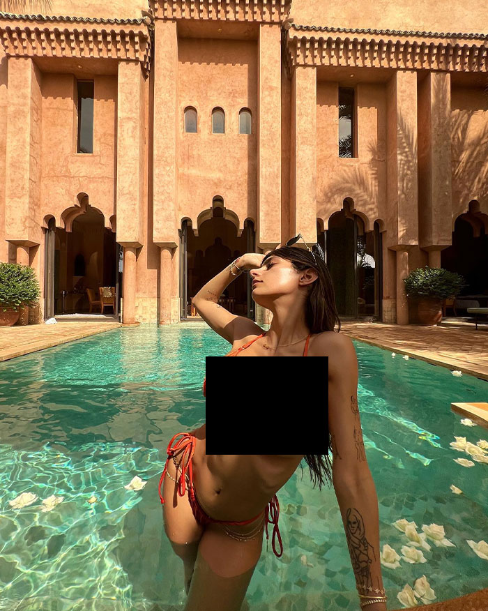 Mia Khalifa in an orange bikini poses in a luxurious pool, leaning back with sunglasses on her head, in front of an ornate Moroccan-style building.