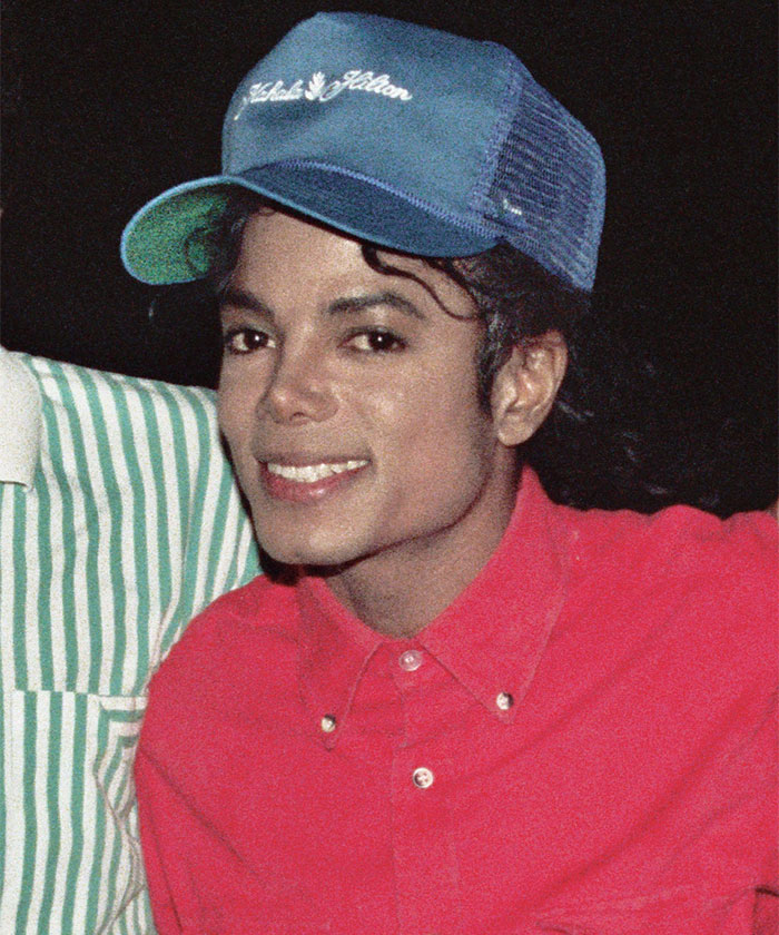 Smiling celebrity in a red shirt and blue cap at an event.