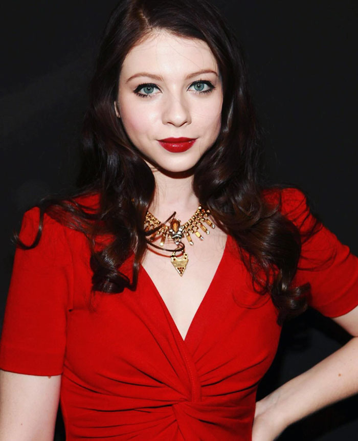 A woman in a red dress with dark hair and bright lipstick, wearing a statement necklace, poses for the camera.