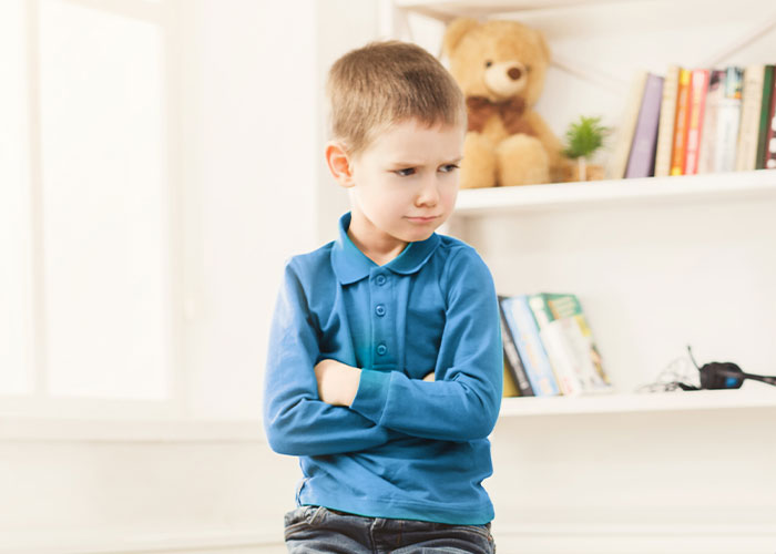 Young boy in a blue shirt crossing arms, standing in a cozy room with a teddy bear and books on a shelf.
