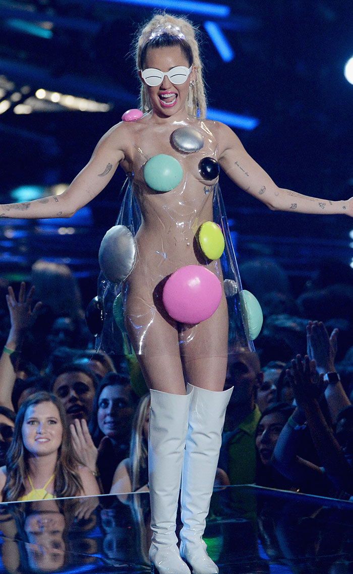 Celebrity in a bold, eccentric outfit with oversized colorful circles and white boots on a stage, sparking conversation.