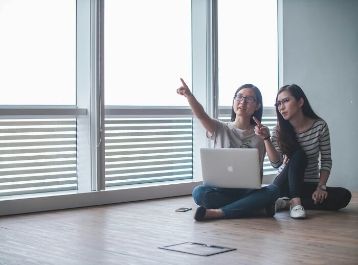 Two women with a laptop sitting on the floor, experiencing culture shocks and discussing something outside the window.
