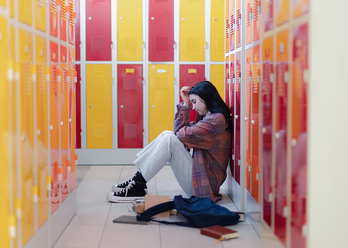 Teen sitting in a school hallway near lockers, looking upset and alone.