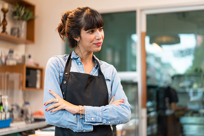 Worker in a denim shirt and apron stands with arms crossed, conveying a thoughtful expression. Worker in a denim shirt and apron stands with arms crossed, conveying a thoughtful expression.