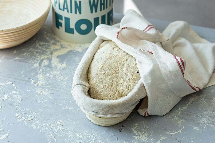 Dough rising in a cloth-covered basket with flour on the table for a minimalism-inspired multi-functional kitchen approach.