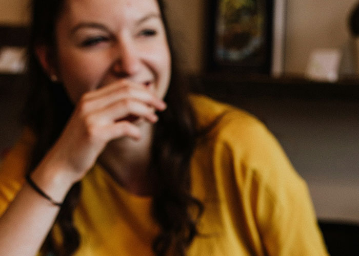Woman in yellow shirt laughing, possibly overhearing something funny in a public setting.