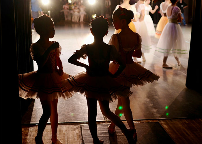 Ballet dancers backstage in silhouette preparing for a performance, with stage lights creating dramatic effects.