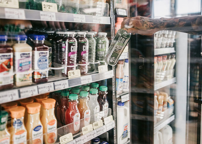 A tattooed arm reaches into a cooler, selecting a green juice bottle among various beverages in a store.