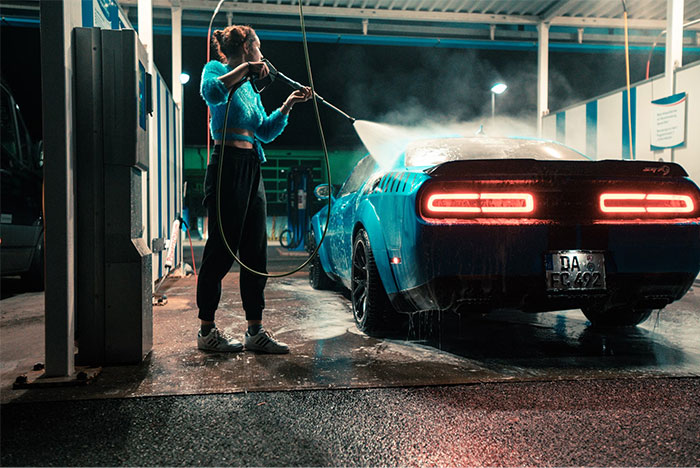 Person washing a blue car at night, using high-pressure hose, showcasing intuition in noticing car details.