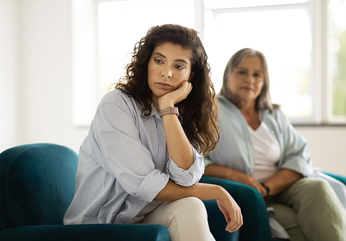 Mother and daughter sitting in a living room, daughter looks upset, reflecting tension. Mother and daughter sitting in a living room, daughter looks upset, reflecting tension.