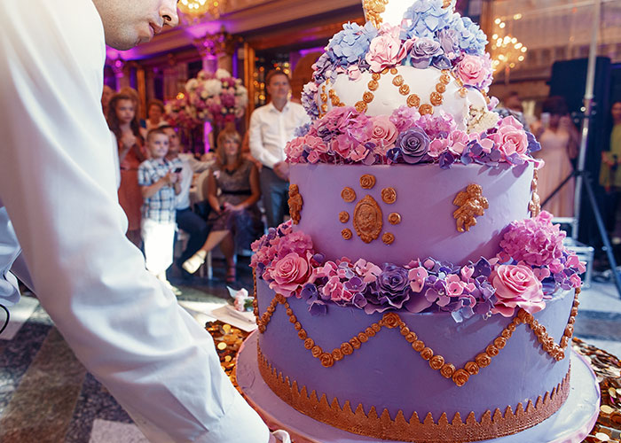 A man adjusts a large purple and pink wedding cake at an event, with onlookers in the background. A man adjusts a large purple and pink wedding cake at an event, with onlookers in the background.