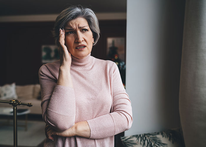 Older woman in a pink sweater looking concerned, with her hand on her face, indoors. Older woman in a pink sweater looking concerned, with her hand on her face, indoors.