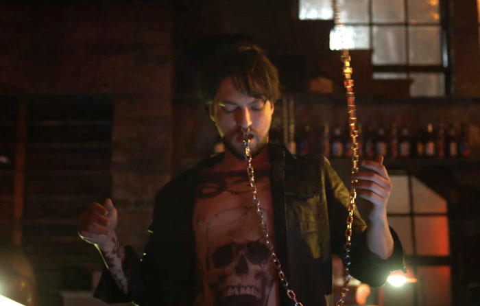 Man from "Final Destination: Bloodlines" in dimly lit room with a skull shirt, performing a trick with chains, related to anticipated movies coming out in 2025.