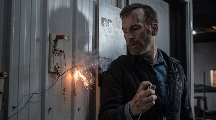 Man from "Nobody 2" holding a sparkler indoors, wearing a denim shirt and jacket, a scene from anticipated movies of 2025.