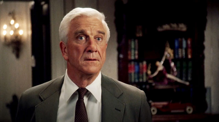 Man from "The Naked Gun" in a suit in a dimly lit room, standing near a shelf with books. 