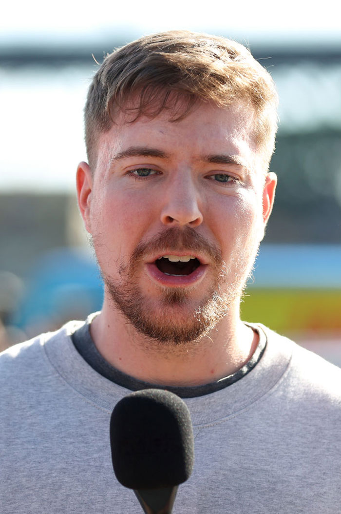 MrBeast with a beard speaking into a microphone outdoors in a casual setting.