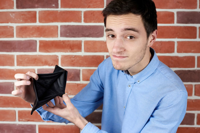 Man in a blue shirt showing an empty wallet, standing against a brick wall, symbolizing neighborhood loans struggle.