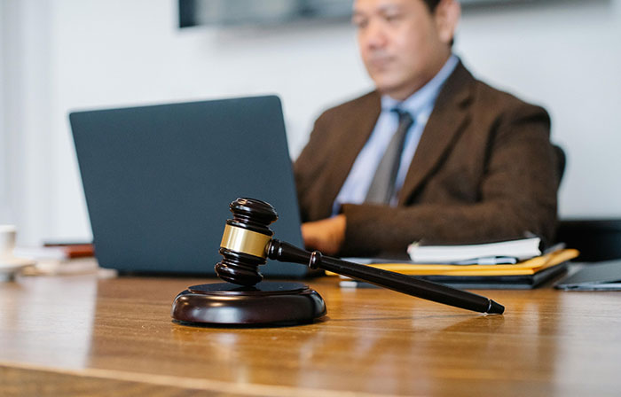 Gavel on a desk with a person working on a laptop in the background, symbolizing decision-making and authority. Gavel on a desk with a person working on a laptop in the background, symbolizing decision-making and authority.