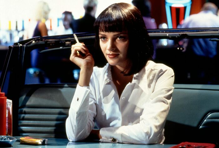 A woman in a 90s-themed setting sitting at a diner, wearing a white shirt and holding a cigarette.