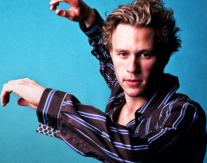 I don't know who this person is, but they're posing with hands raised against a blue background wearing a striped shirt.