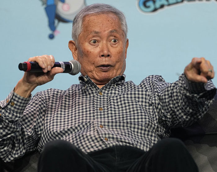 Man holding a microphone with an animated expression, wearing a checkered shirt, at a public event.