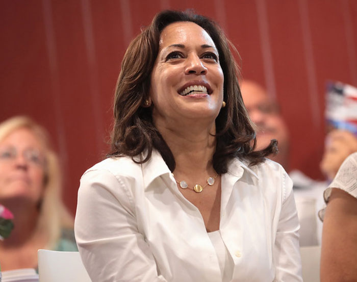 A person in a white blouse smiling warmly in a relaxed indoor setting, surrounded by a group of people.