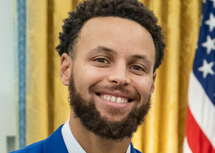 Smiling man in a blue jacket stands in a room with golden drapes.