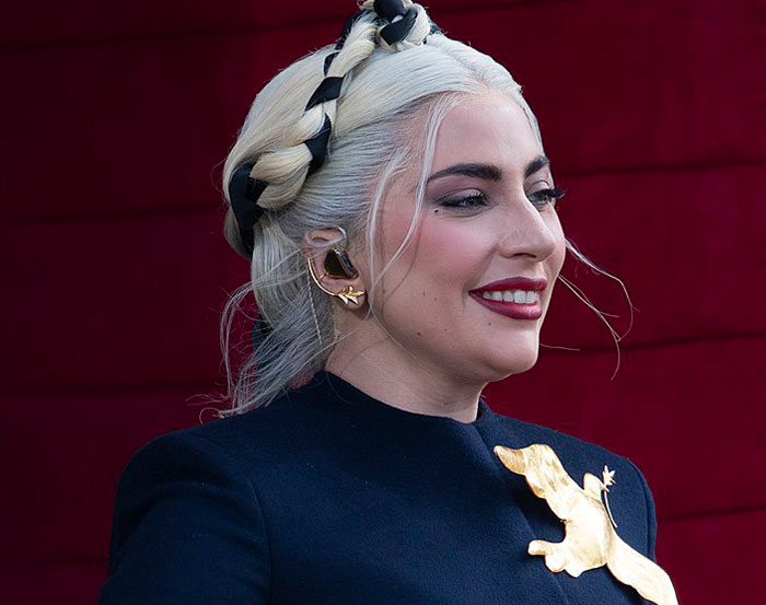 A person with blonde hair and a braided headband, smiling and wearing a dark outfit with gold embellishment.