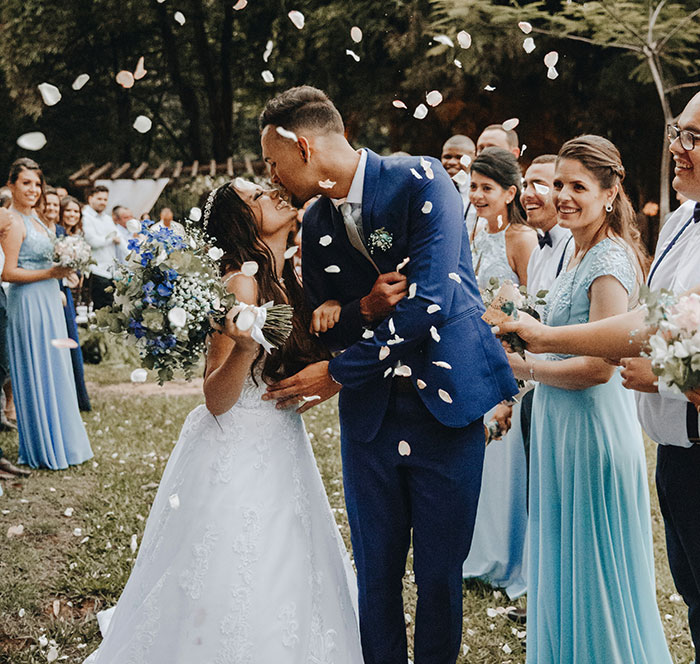 Wedding couple kissing in backyard ceremony, surrounded by friends in blue dresses, amid scattered flower petals. Wedding couple kissing in backyard ceremony, surrounded by friends in blue dresses, amid scattered flower petals.