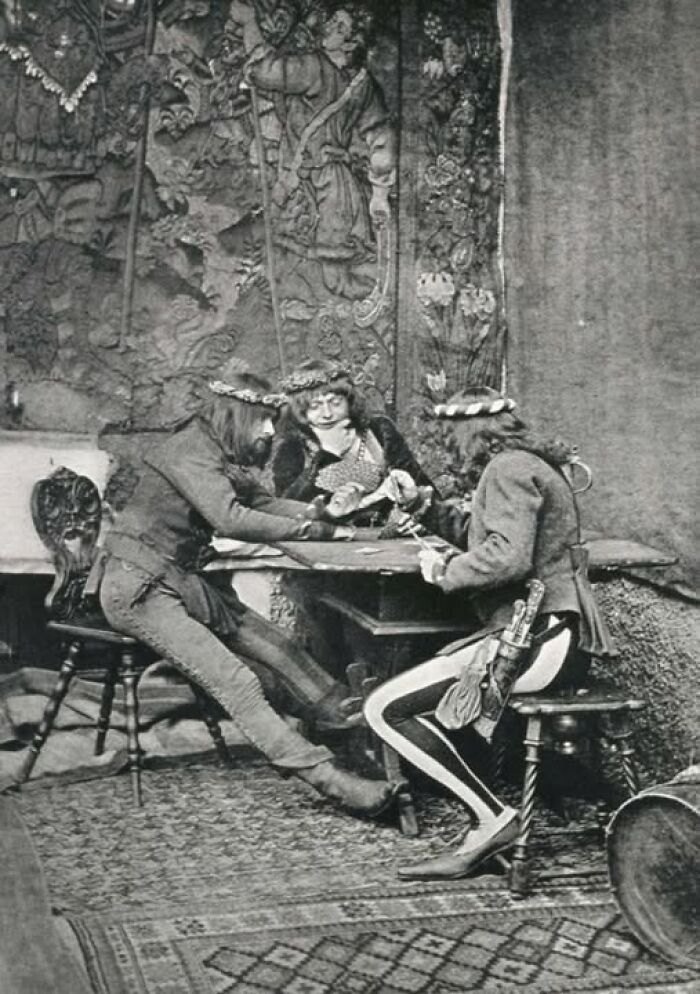 Historical scene with three people in vintage attire at a table, showing intricate tapestry in the background.