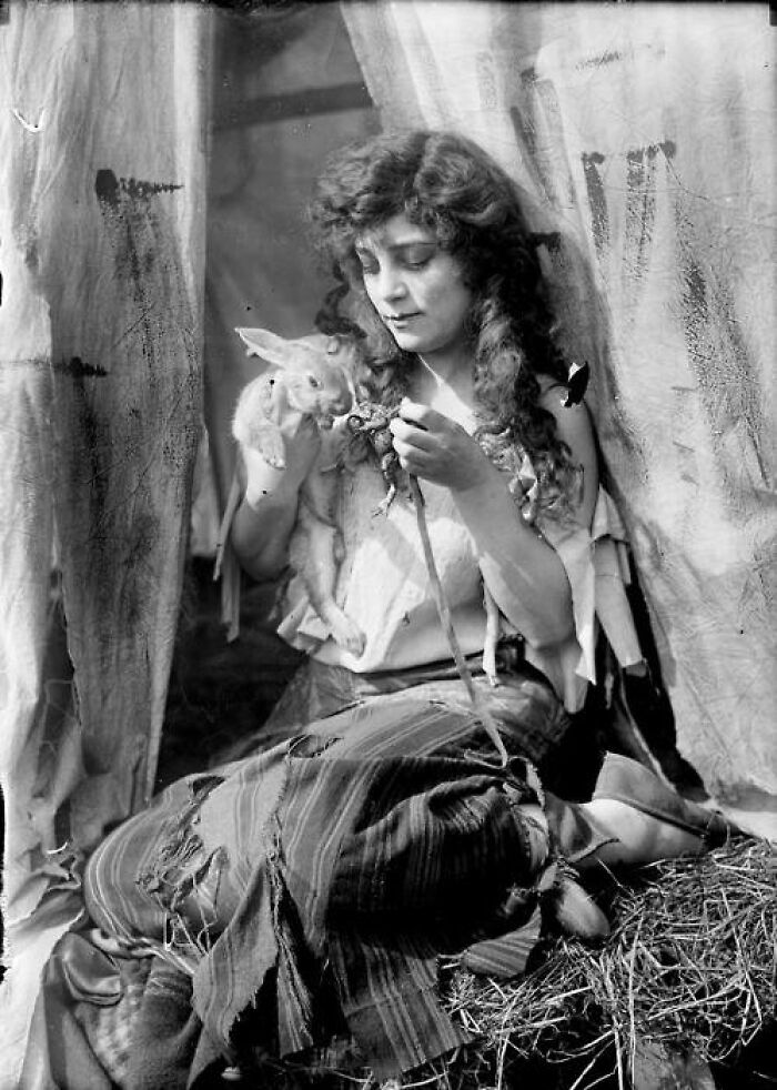 A woman from a century ago, sitting with a rabbit, wearing period clothing, in a rustic setting.