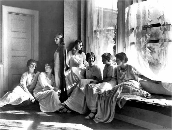 Women in vintage dresses gathered by a sunlit window, embodying a timeless resonance from a hundred years ago.