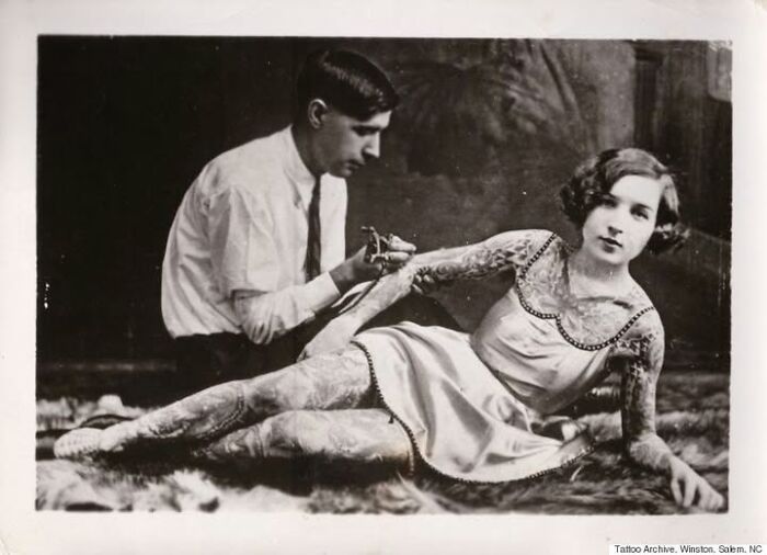 Tattoo artist working on a woman's arm in an early 20th-century setting, showcasing historical tattoo practices.