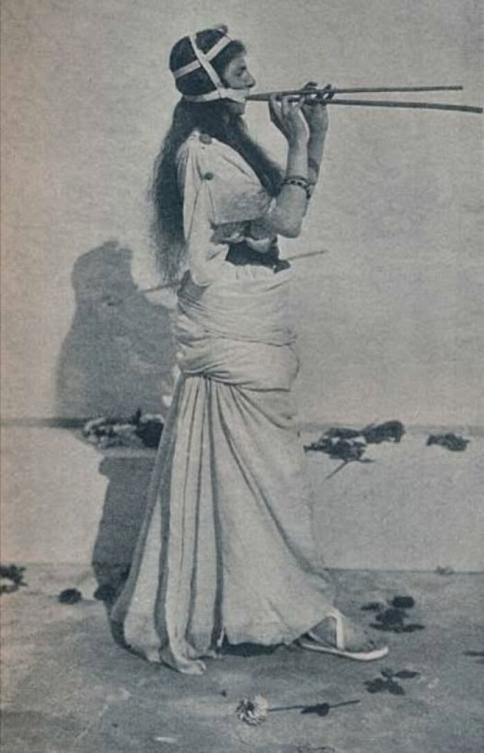 A woman in historical attire plays a long flute, showcasing cultural resonance from a hundred years ago.