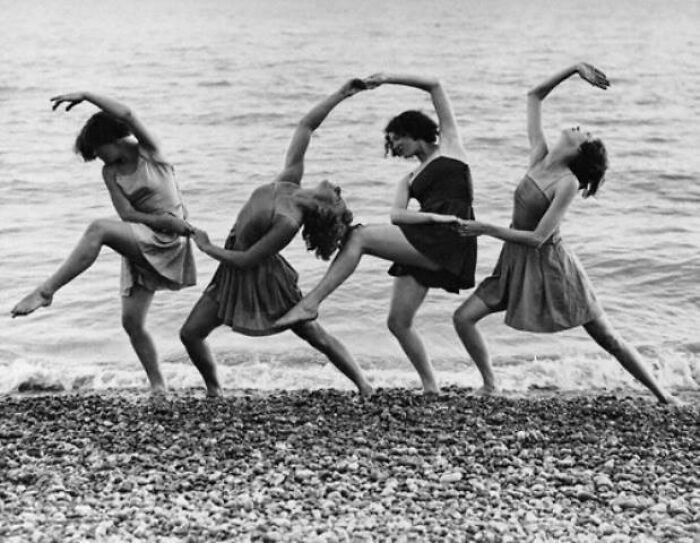 Four women in vintage dresses perform a dance on a rocky shoreline; a photo resonating through the years.