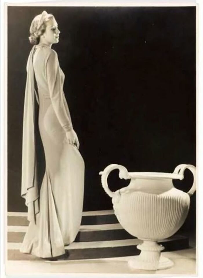 Woman in vintage gown standing on stairs beside ornate vase in historical photograph.