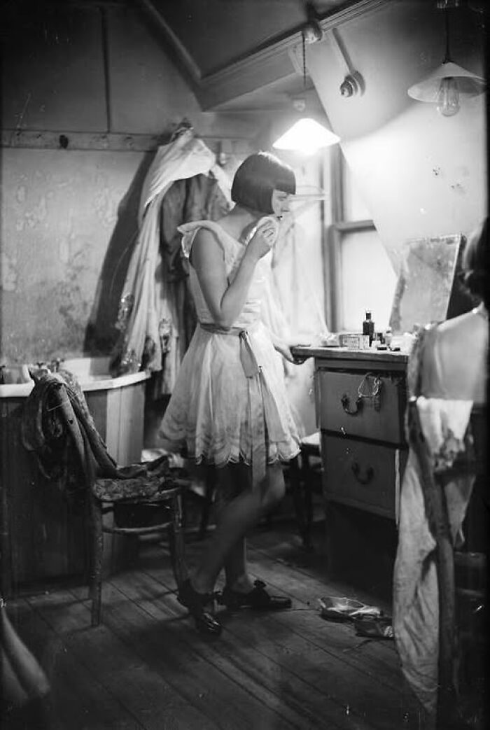 Woman in vintage dress applying makeup in a dimly lit room, capturing an evocative moment from over a hundred years ago.