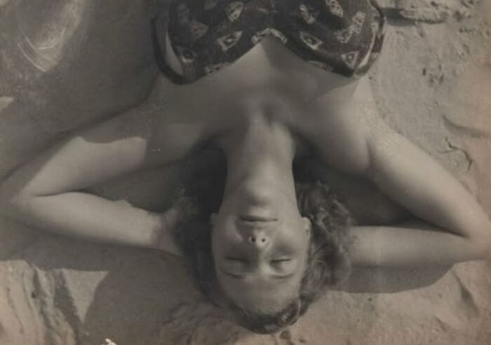 Woman lying on sand with arms behind her head, wearing a patterned swimsuit, from a hundred years ago.