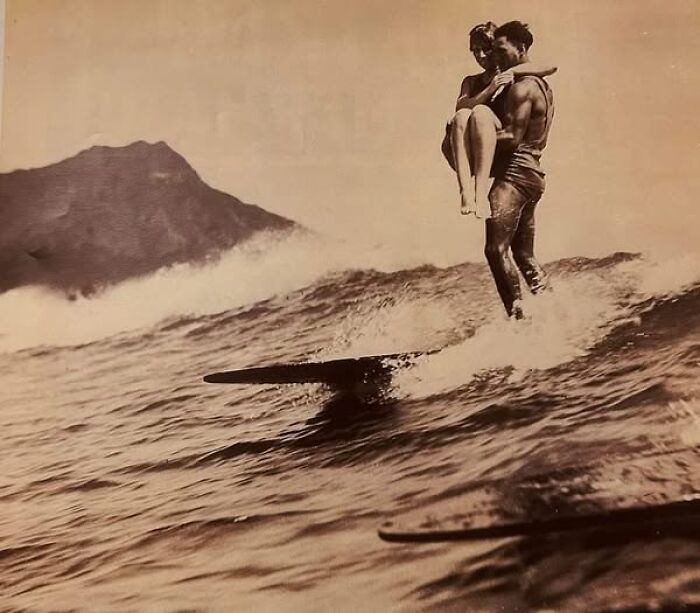 Vintage photo of a man carrying a woman while surfing, showcasing moments from a hundred years ago that continue to resonate today.