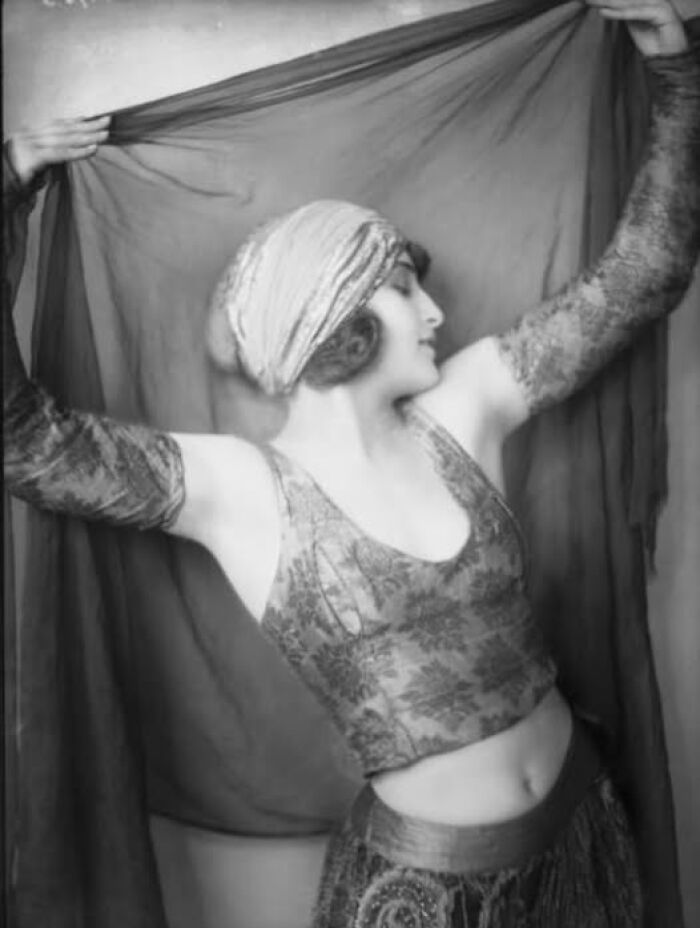 Dancer in vintage attire striking a pose with elegant arm movement, showcasing fashion from over a hundred years ago.