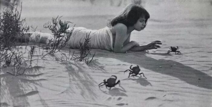 A vintage photo of a woman lying on sand, observing crabs, evokes timeless interest.